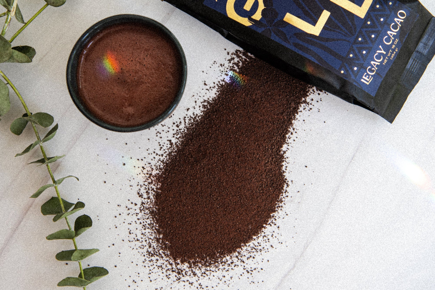 Amazonian Gold Legacy Cacao packaging with cocoa powder on a light surface with eucalyptus leaves.